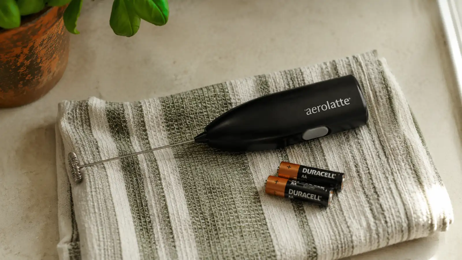 Close-up of a worn black Zulay milk frother lying on a kitchen towel next to two spent AA batteries